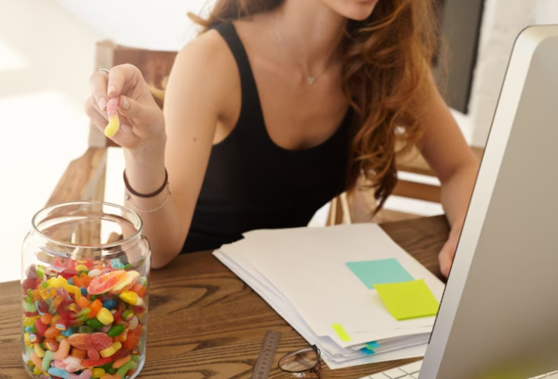 caucasian female office worker taking marmalade from the big jar at work. the girl eats candies for improvement of work of a brain during hard work over the financial statement