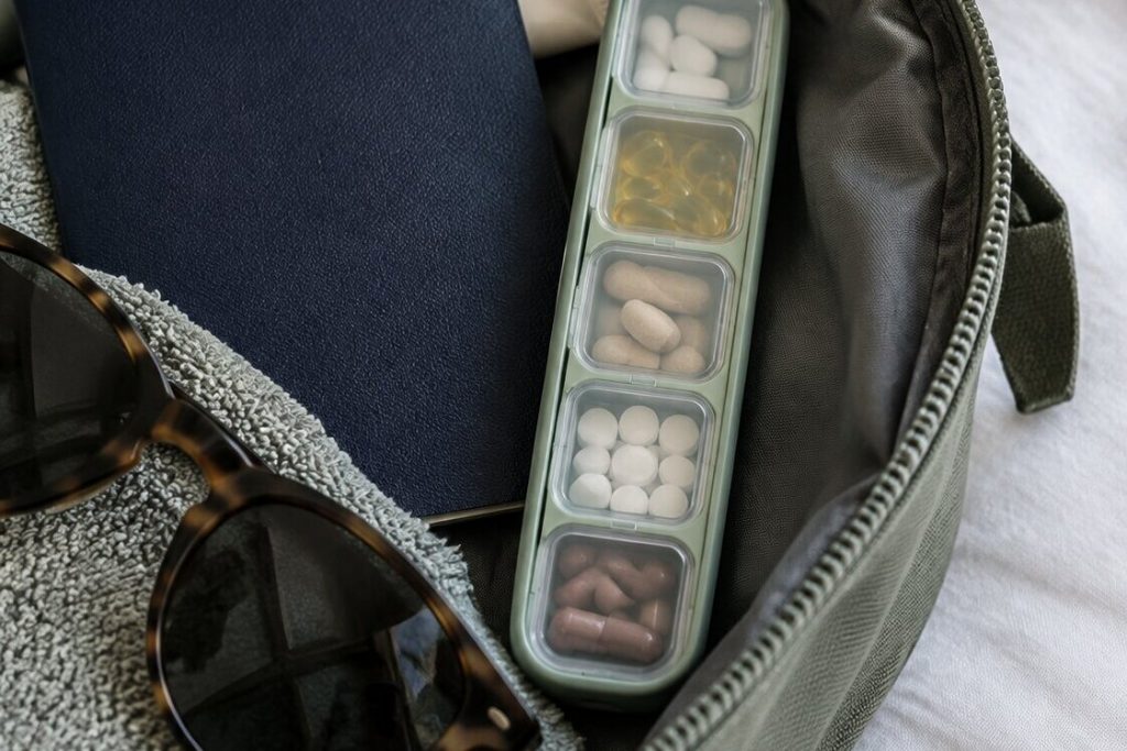 A slim travel pill case with five clear compartments of vitamins and capsules in an open toiletry bag next to a passport and sunglasses