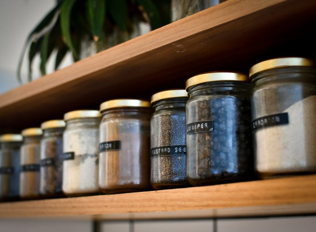 A row of labeled glass spice jars with gold lids on a wooden shelf