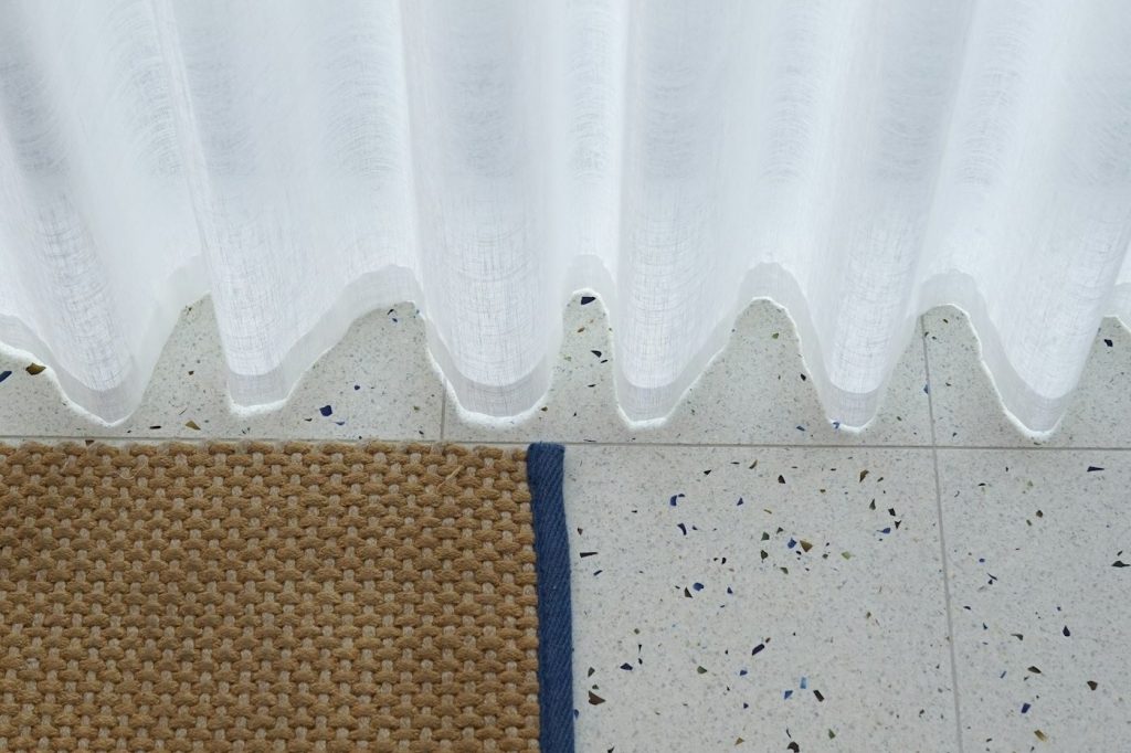 A woven jute bath mat with a blue border on a speckled terrazzo floor below a sheer white curtain