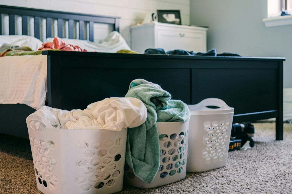 Three white plastic laundry baskets piled with towels and clothes on the carpet at the foot of a bed