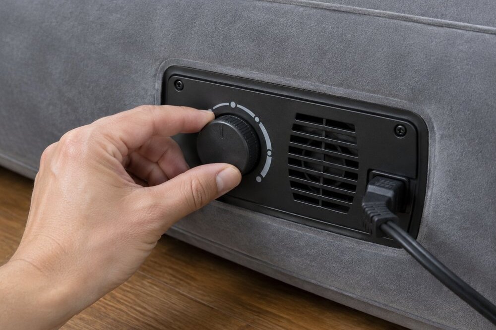 A hand turning the dial on the built-in electric pump of a gray flocked air mattress
