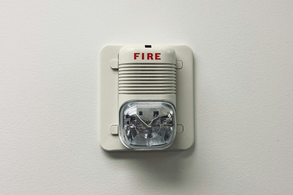 A white wall-mounted fire alarm with a strobe light on a plain white wall