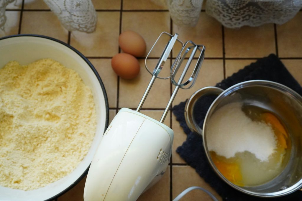 Hand mixer beside bowls of flour, sugar, and eggs on a tiled counter