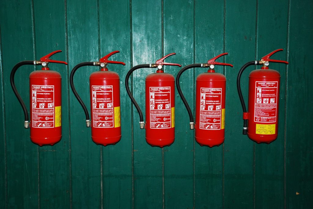 Five red fire extinguishers with black hoses mounted in a row on a green wooden wall