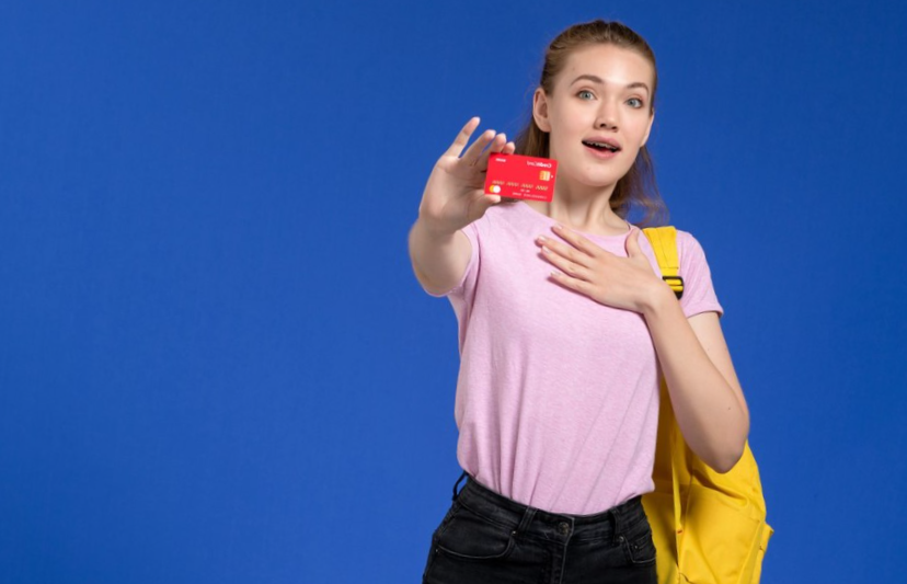 front view of young female in pink t-shirt wearing yellow backpack holding plastic red card

