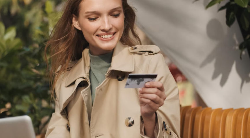 Beautiful smiling girl dressed in trench coat holding credit card and smiling working on laptop in cafe outdoor modern technology