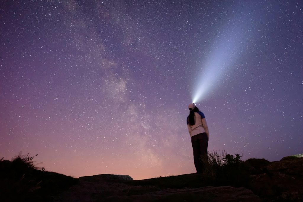 A person on a hilltop points a headlamp beam up into a starry purple sky