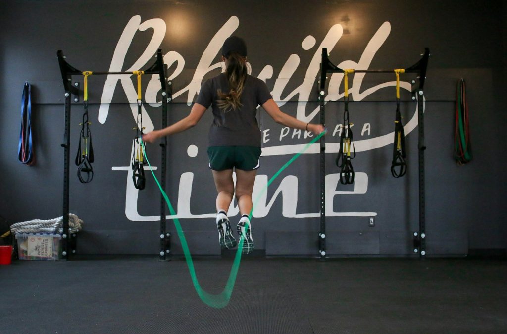 A woman jumping rope in a gym with a green rope, in front of wall-mounted training racks
