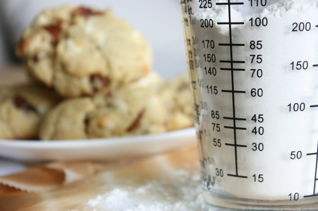 A glass liquid measuring cup with printed markings next to a plate of cookies