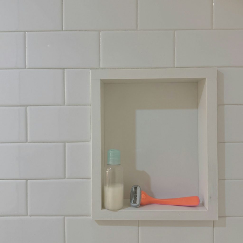 A small recessed tile niche in a white shower wall holding a bottle and a razor