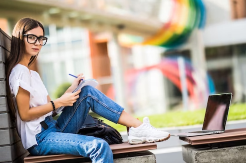 female student taking notes seated on a bench in a park in front of her laptop