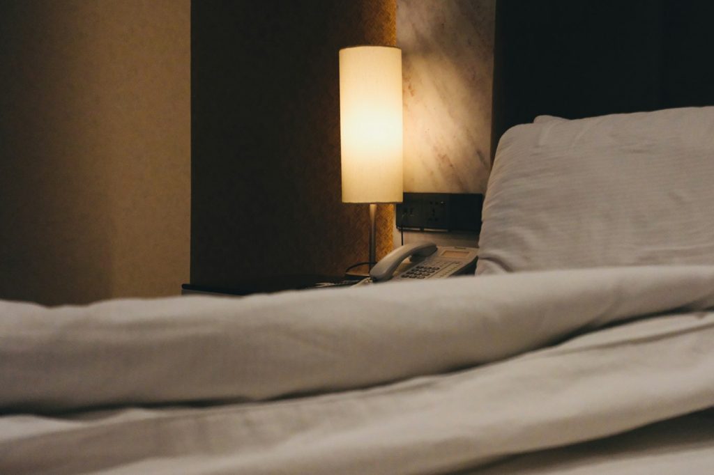 A dim bedside lamp glowing beside a bed with rumpled white sheets