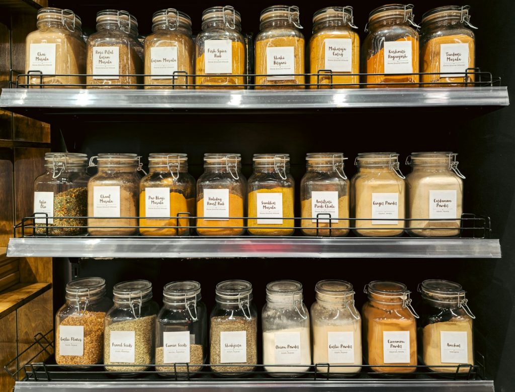 Three metal shelves lined with glass jars of labeled seasonings
