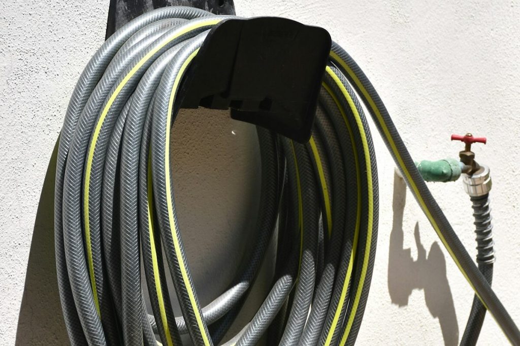 A gray and yellow garden hose coiled on a black wall-mounted hanger next to an outdoor spigot