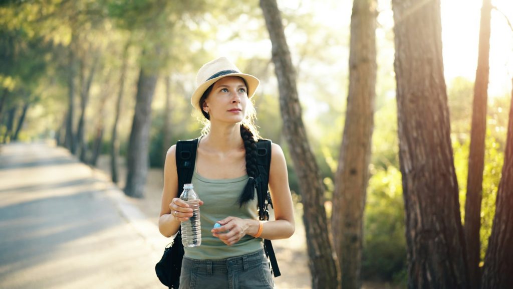 A woman in a sun hat carrying a backpack and a water bottle on a tree-lined path