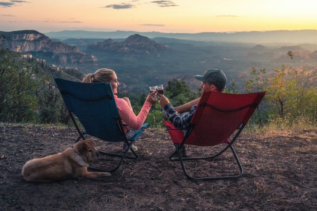 Two people in folding camping chairs with a dog, overlooking a canyon at sunset