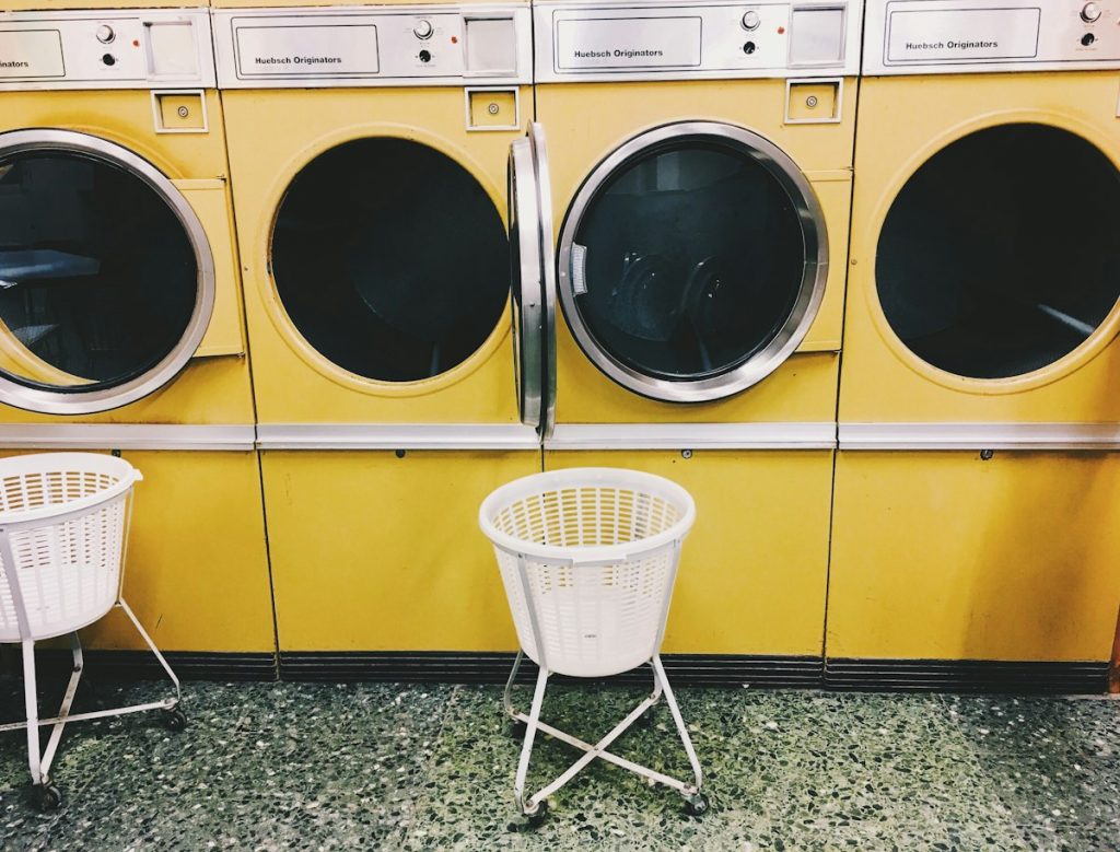 A white rolling laundry basket on a metal stand in front of a row of yellow front-load dryers at a laundromat