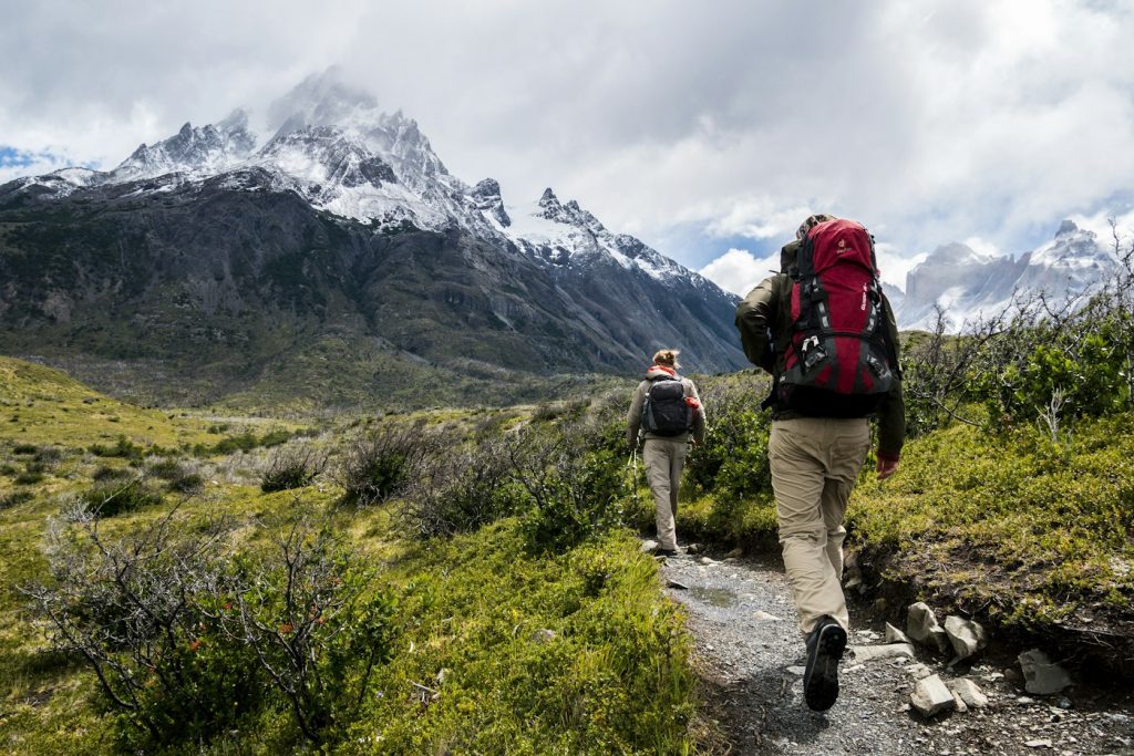 Two hikers with backpacks on a mountain trail below snowy peaks