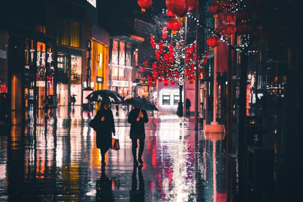 Two people with umbrellas walking down a wet, reflective street lined with red lanterns at night