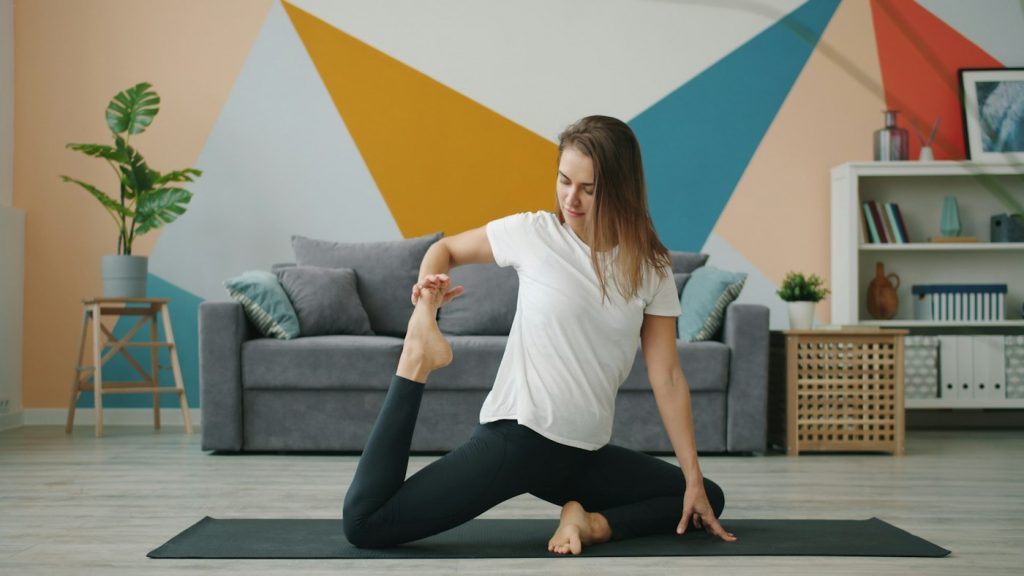 A woman stretching on a dark yoga mat in a bright living room with a gray sofa
