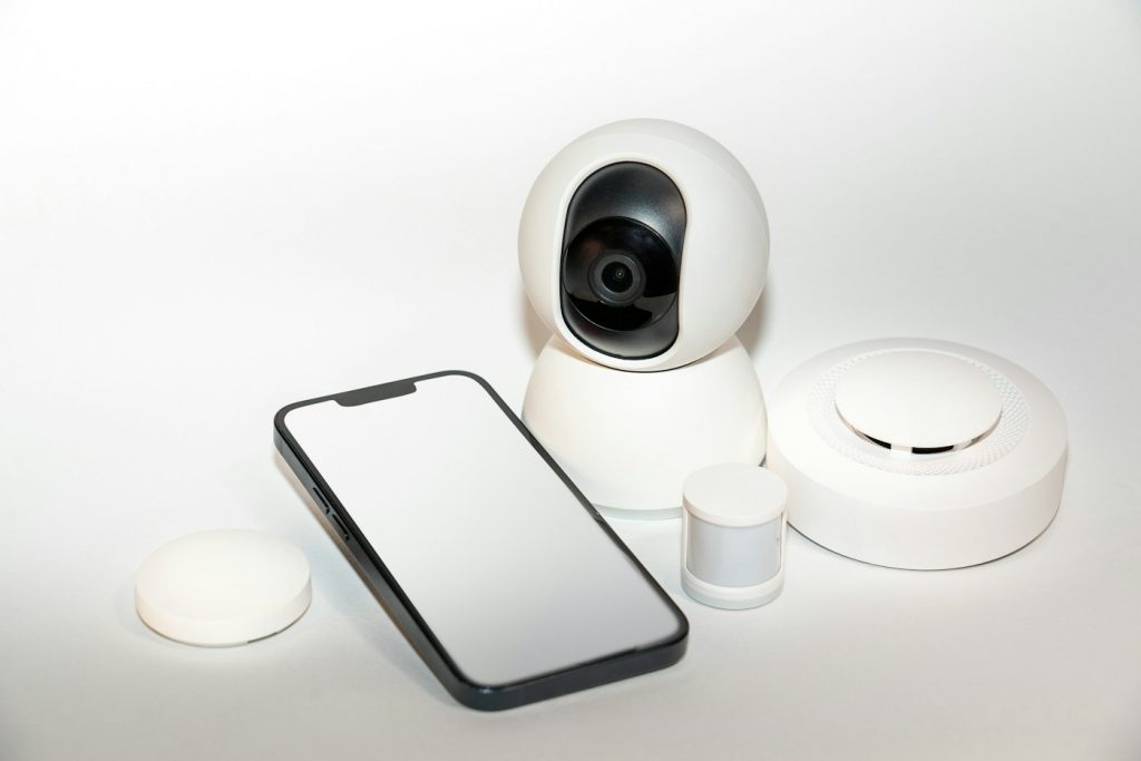 White baby monitor camera next to a smartphone and small smart home sensors