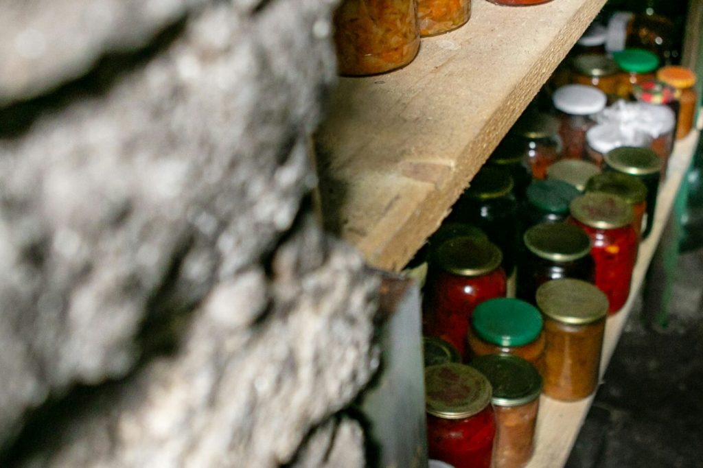 Rows of home-preserved jars in red, orange, and green on wooden shelves in a stone cellar