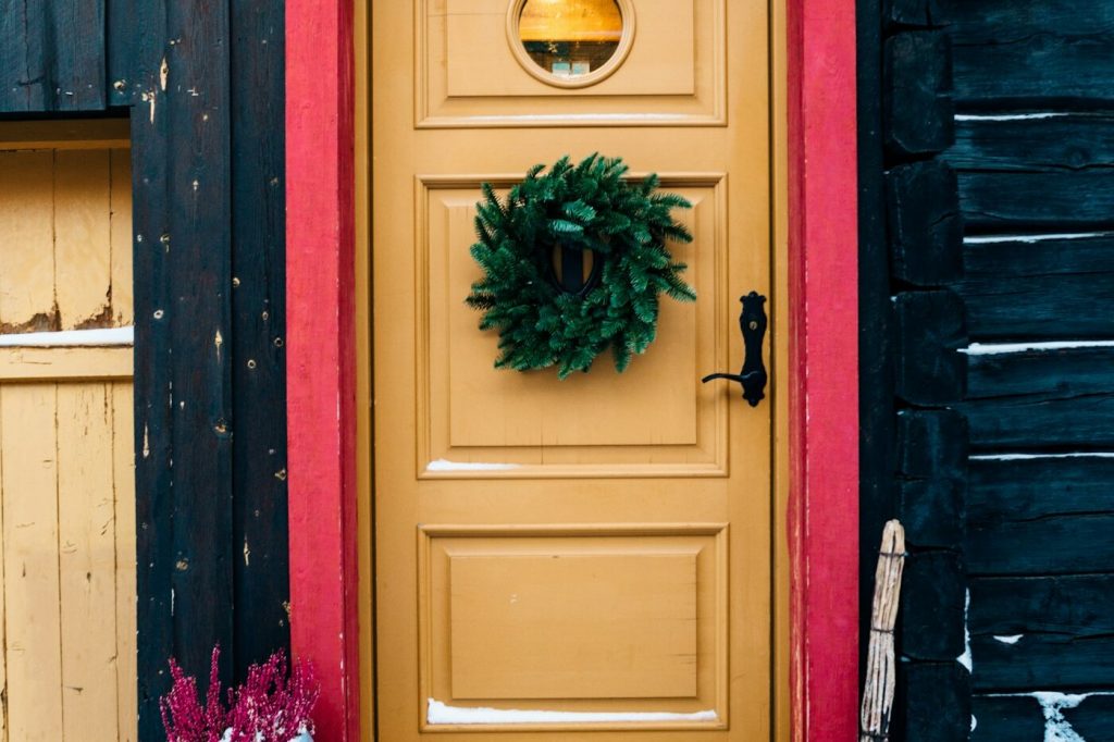 A mustard yellow front door with a green wreath, framed in red trim, with snow on the step