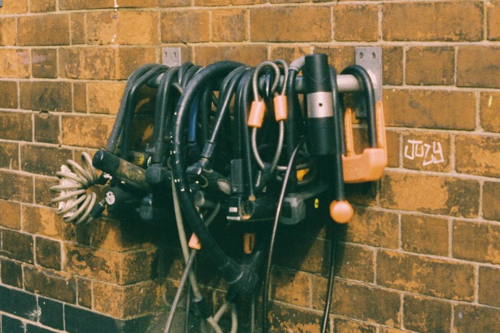 A cluster of bike locks, cables, and chains hanging from hooks on a brick wall under a bike parking sign
