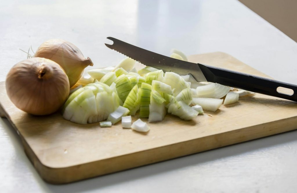 Diced onion and a whole onion beside a serrated knife on a light wooden cutting board