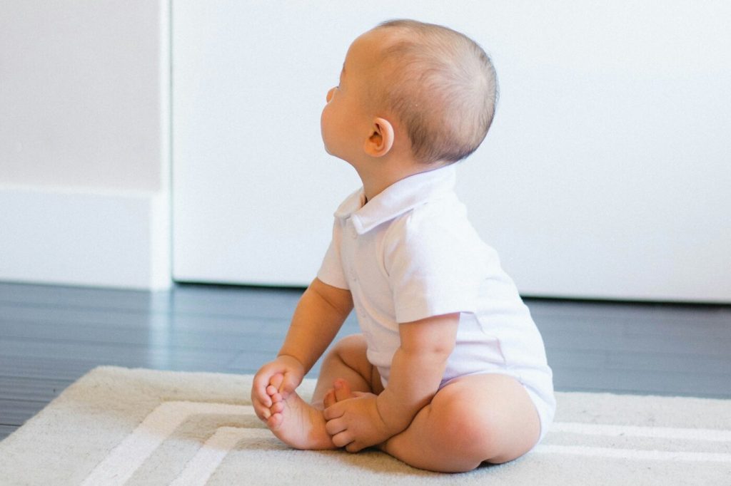 A baby in a white bodysuit sitting on a beige rug and looking up at a wall