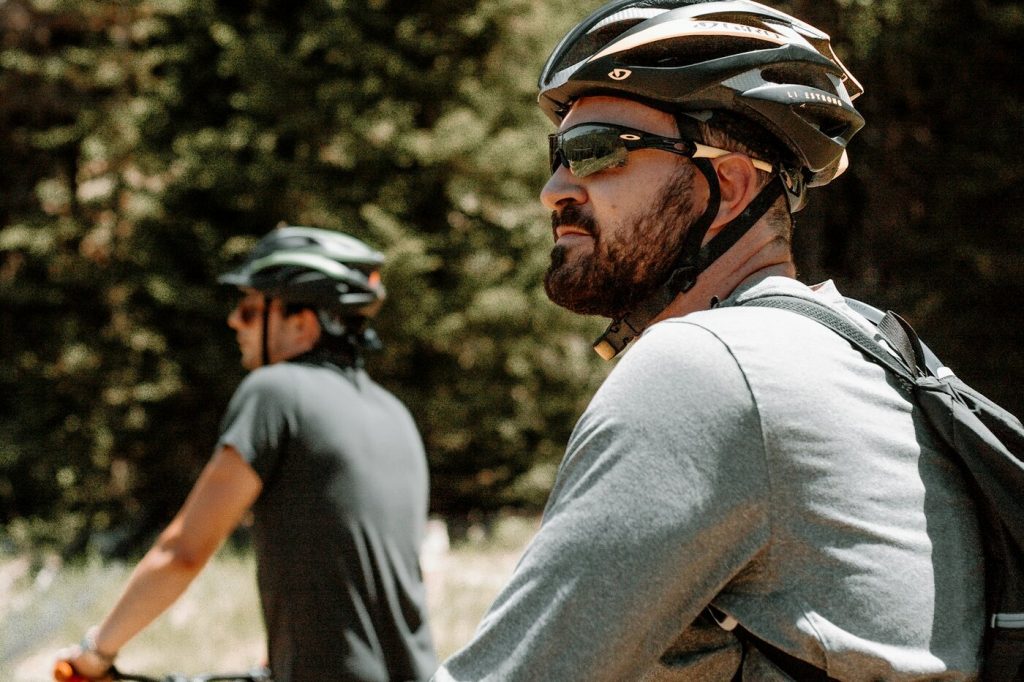 Two mountain bikers wearing vented bike helmets and sunglasses pause on a trail beside a pine forest