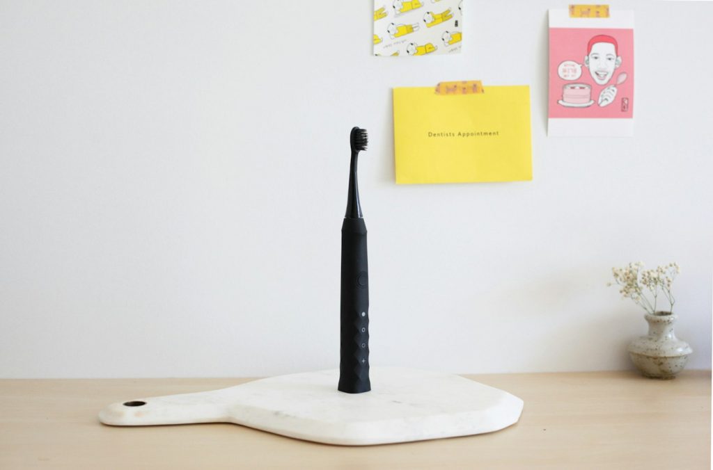 A black electric toothbrush standing upright on a white stand against a light wall