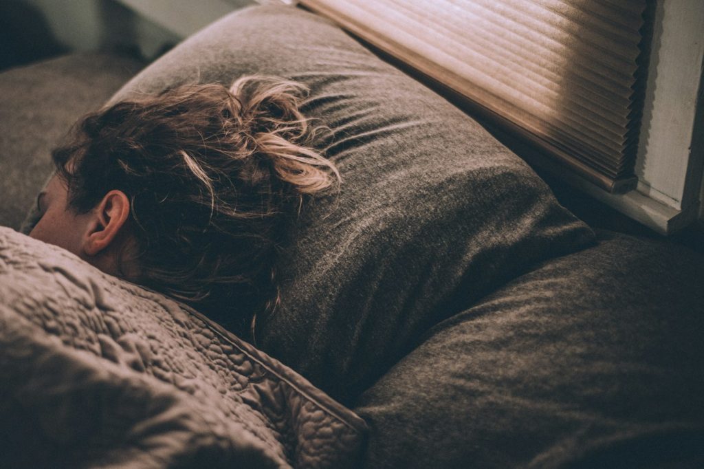 A person asleep face down on a gray pillow next to a window with half-open blinds