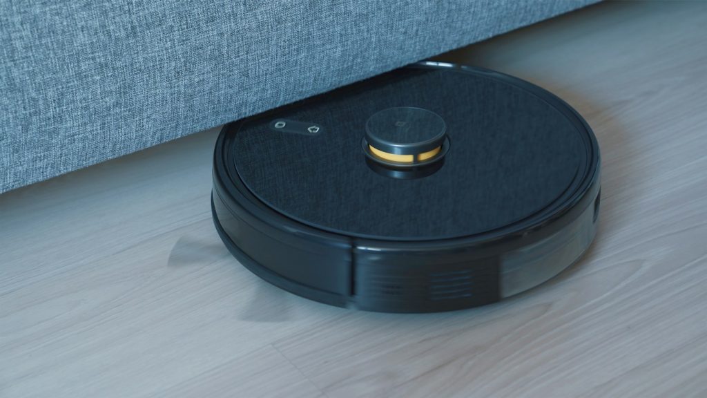 Black robot vacuum cleaning a wooden floor beside a sofa