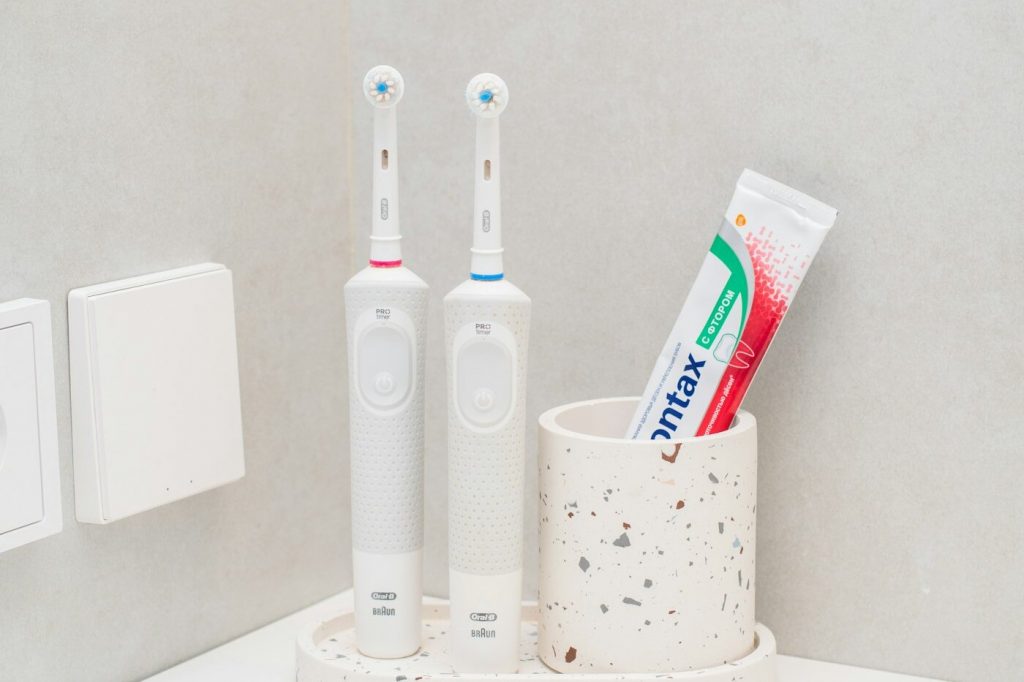 Two white electric toothbrushes and a tube of toothpaste in a holder beside a bathroom sink