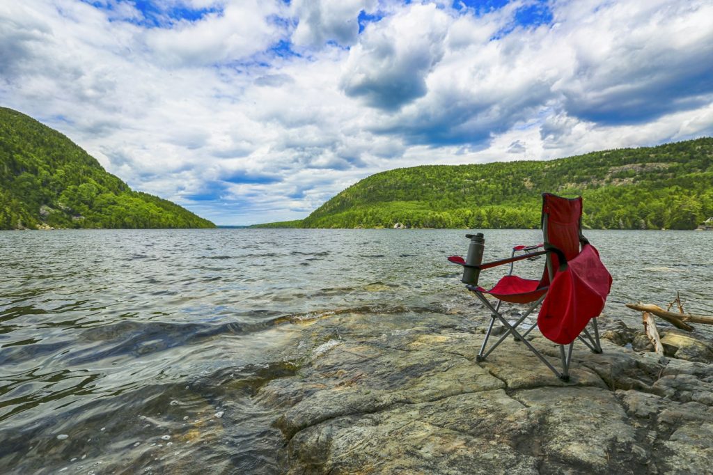 Red folding camping chair on a rocky lakeshore with green hills