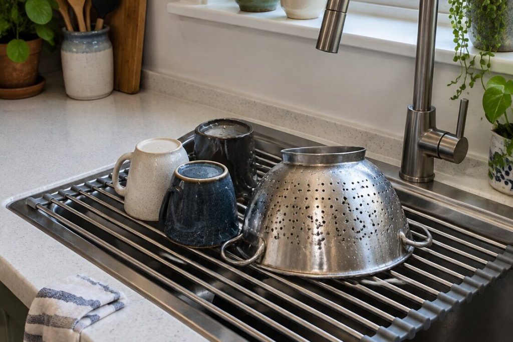 A roll-up stainless steel and silicone dish drying rack laid across a kitchen sink holding mugs and a metal colander