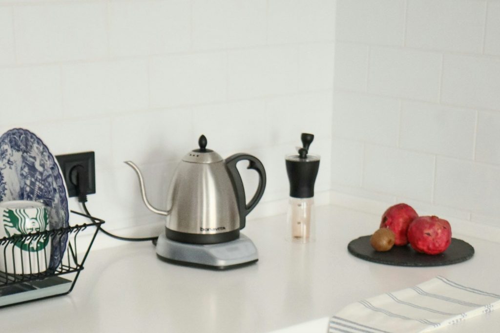 A stainless steel gooseneck electric kettle on its base on a white kitchen counter, next to a coffee grinder and a plate of fruit