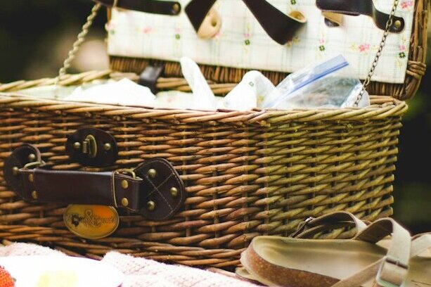 An open wicker picnic basket with leather straps sitting on a soft pink blanket