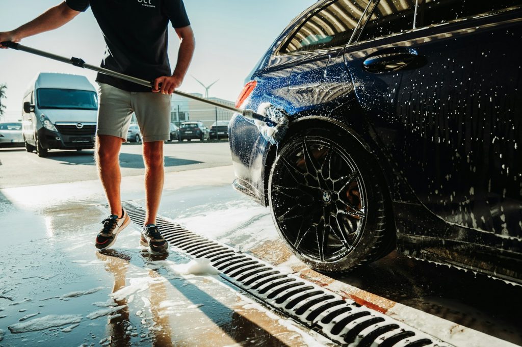 Person using a pressure washer wand to rinse a dark car on a wet driveway