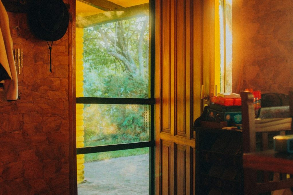 A sunlit open doorway with a glass screen door leading from a warm tiled entry hall to a covered porch