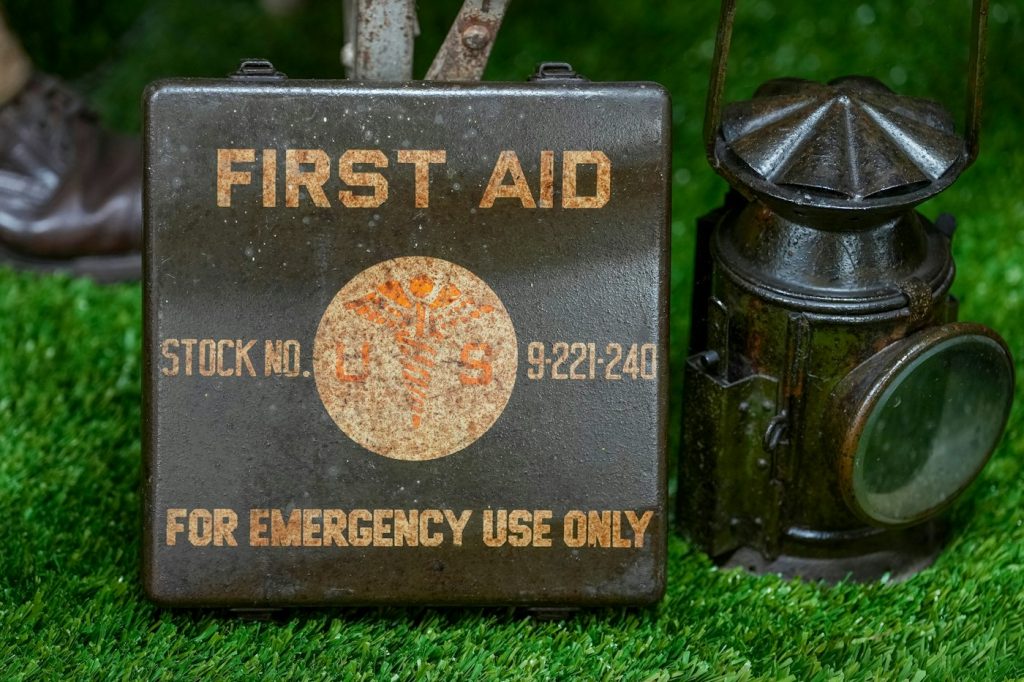 A vintage metal first aid box marked for emergency use next to an old lantern on grass