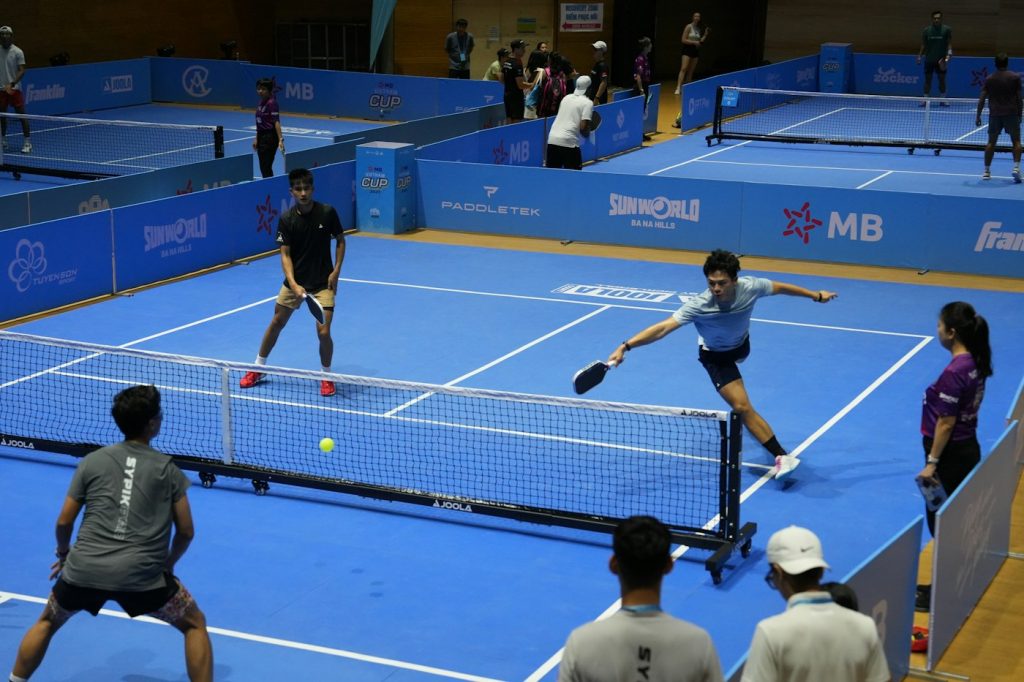 Four players in a doubles pickleball match on a blue indoor court
