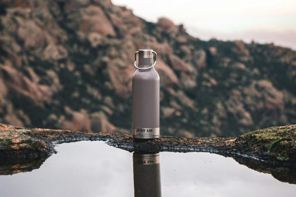 A metal water bottle standing on a rock beside a still pool reflecting the sky